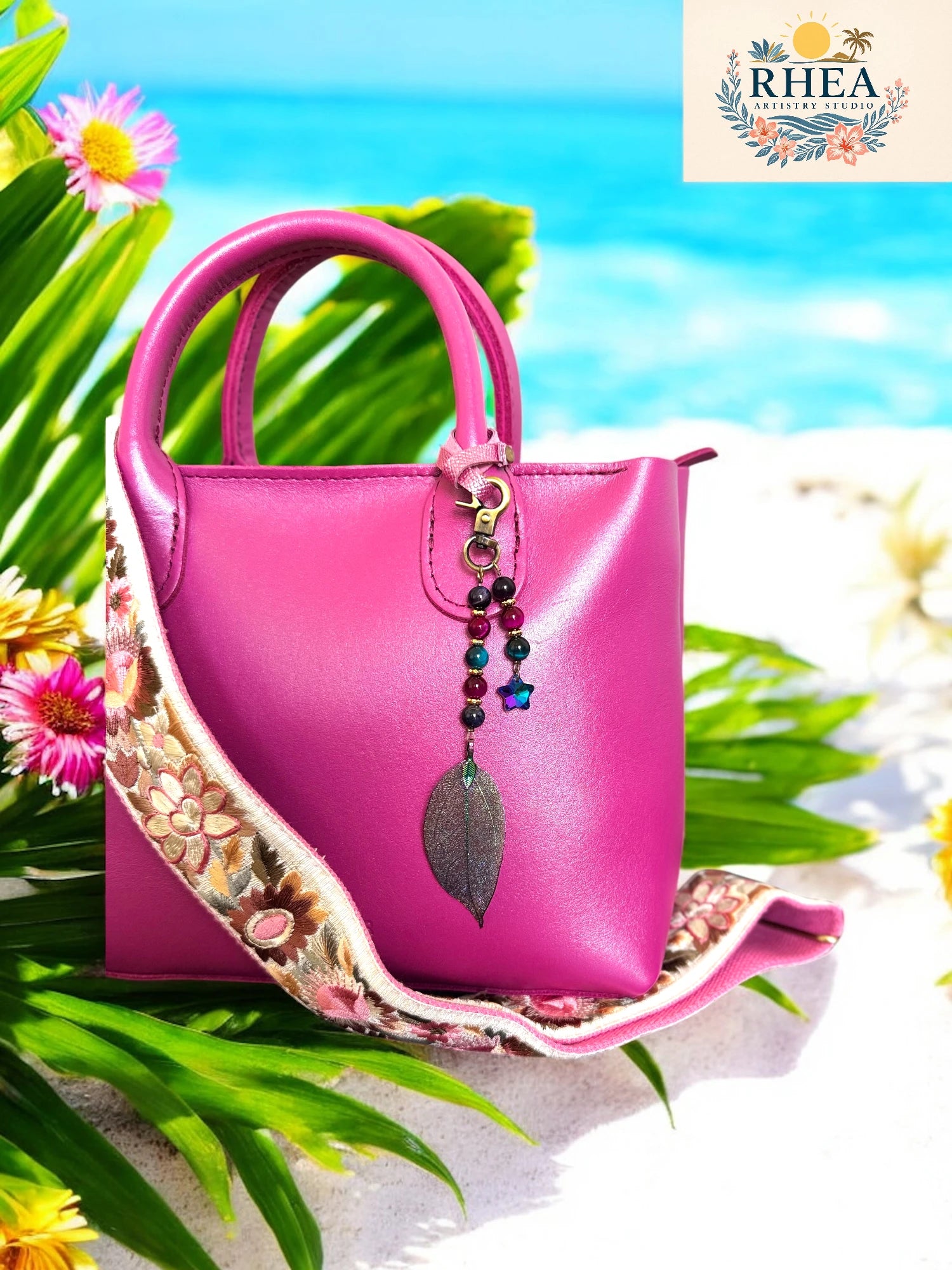 Pink handbag with decorative strap on a beach background, featuring the RHEA brand.