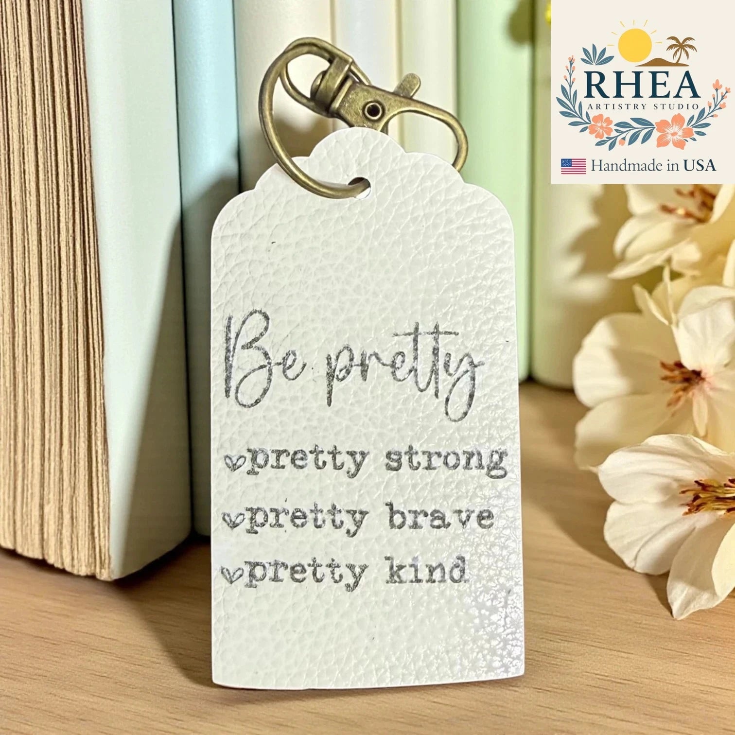 Motivational leather tag with 'Be pretty, pretty strong, pretty brave, pretty kind' text on a wooden surface with books and flowers.
