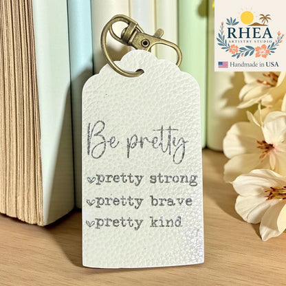 Motivational leather tag with 'Be pretty, pretty strong, pretty brave, pretty kind' text on a wooden surface with books and flowers.