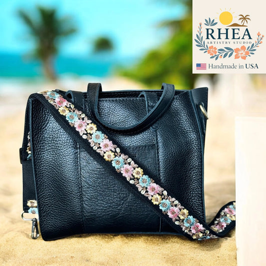 Black leather handbag with a floral strap on a beach background, featuring the RHEA brand.
