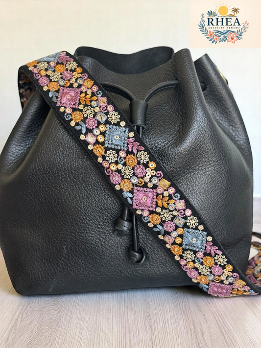 2-inch strap with bold embroidered lavender, gold, and blue squares surrounded by florals on a black base, styled with a black leather bag on a wood countertop