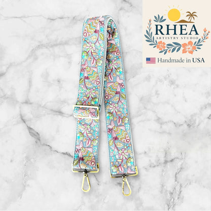 Floral patterned purse strap with gold hardware on a marble background, featuring RHEA Artistry Studio branding.