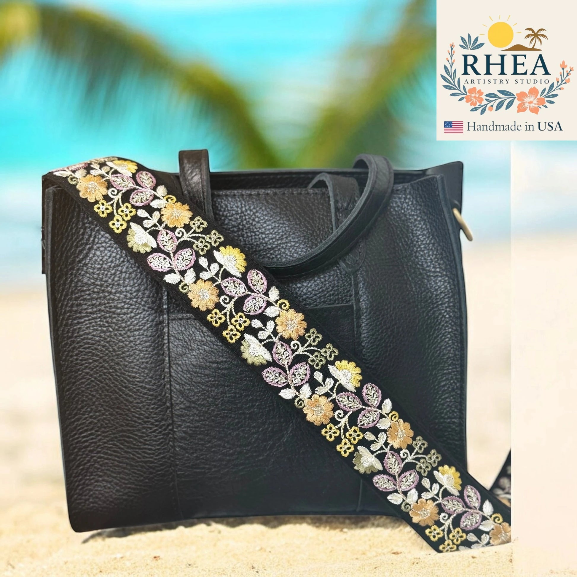 Black handbag with a floral strap on a beach, featuring the RHEA brand.