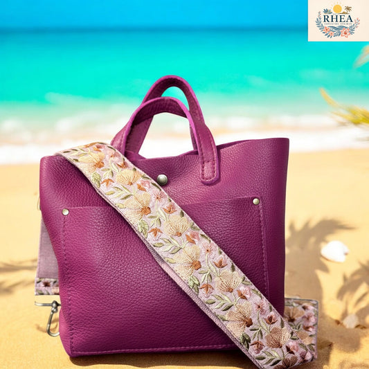 A detailed floral embroidered bag strap in earthy tones displayed on a purple-toned leather tote. Set against a sunny beach backdrop, the strap showcases soft petals, leaf details, and layered textures.