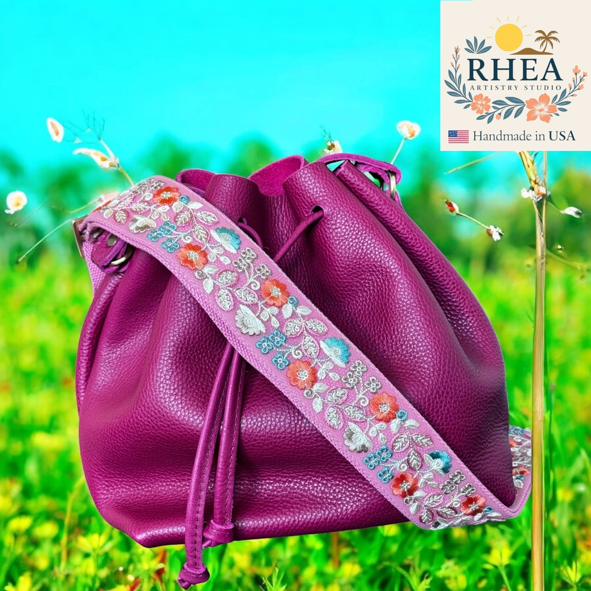 Purple leather bag with floral strap on a grassy background, featuring the RHEA brand.