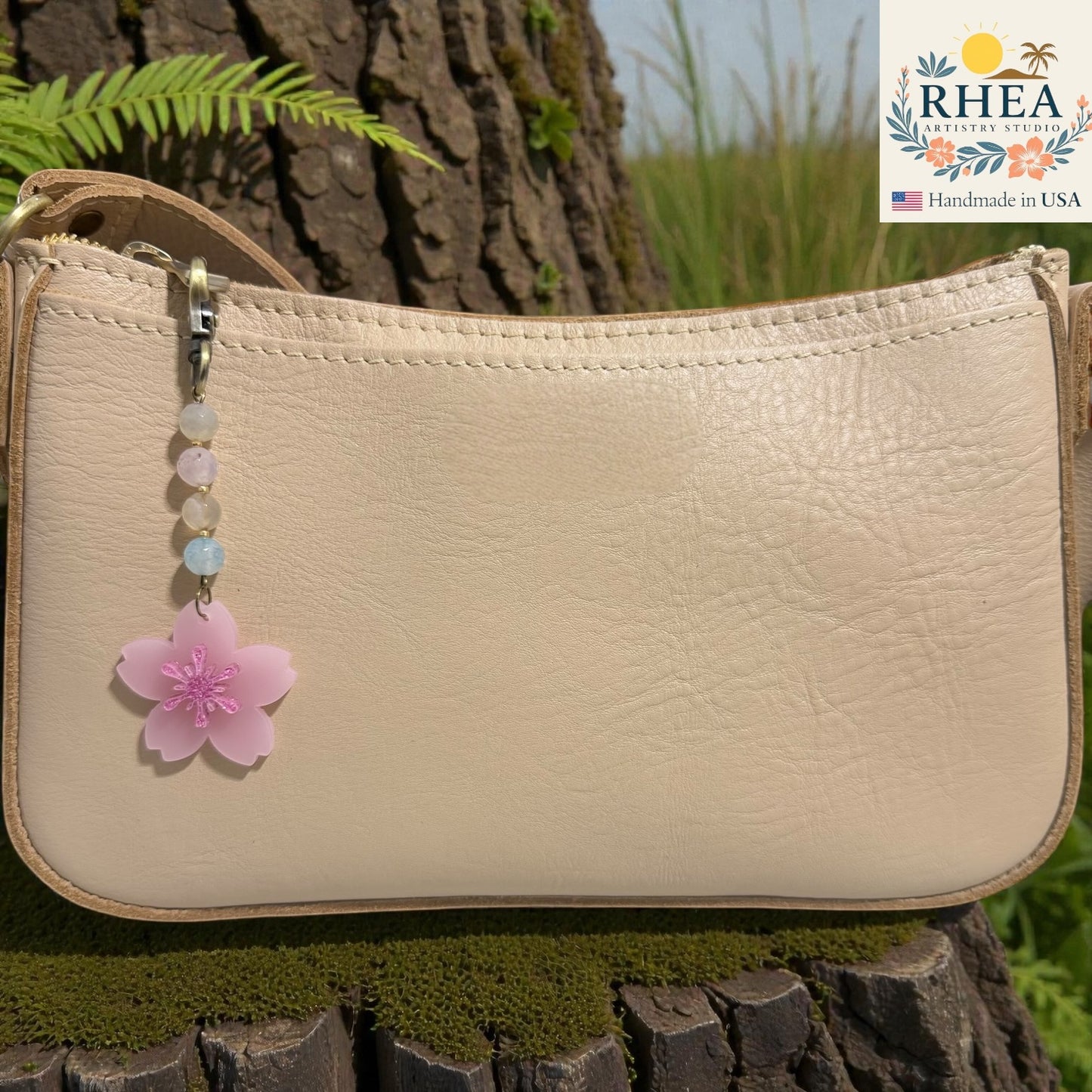 Beige handbag with a pink flower charm on a wooden surface, featuring the RHEEA brand. Frosted cherry blossom acrylic bag charm clipped to neutral handbag