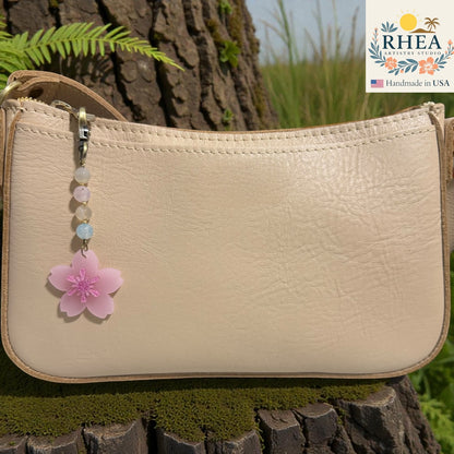 Beige handbag with a pink flower charm on a wooden surface, featuring the RHEEA brand. Frosted cherry blossom acrylic bag charm clipped to neutral handbag