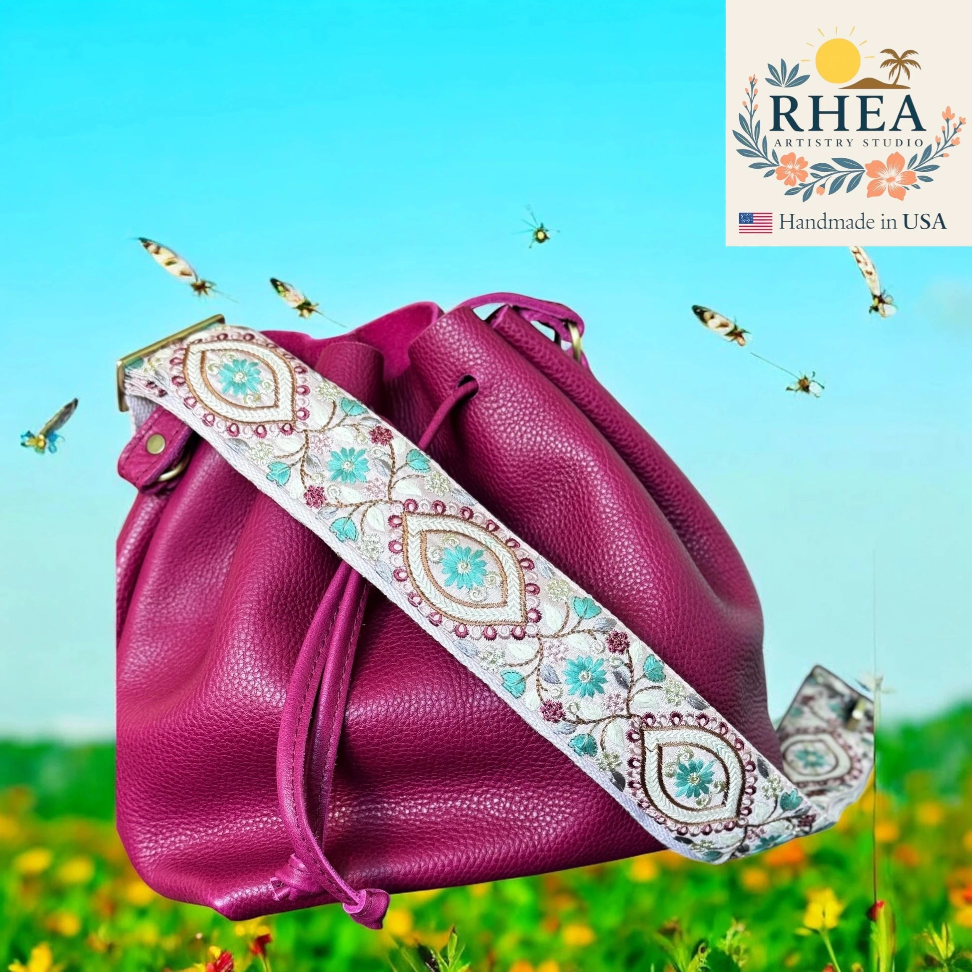 Pink leather bag with an embroidered strap on a scenic background, featuring the RHEA brand.