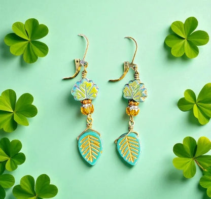 Pair of leaf-shaped earrings with gold and turquoise details on a light green background with shamrock leaves.