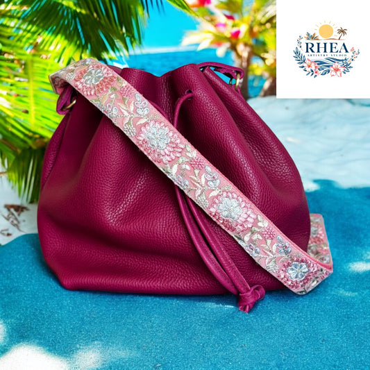 A dreamy strap with silvery white and soft pink florals embroidered on a pale pink webbing base, styled with a magenta leather bag at beach

