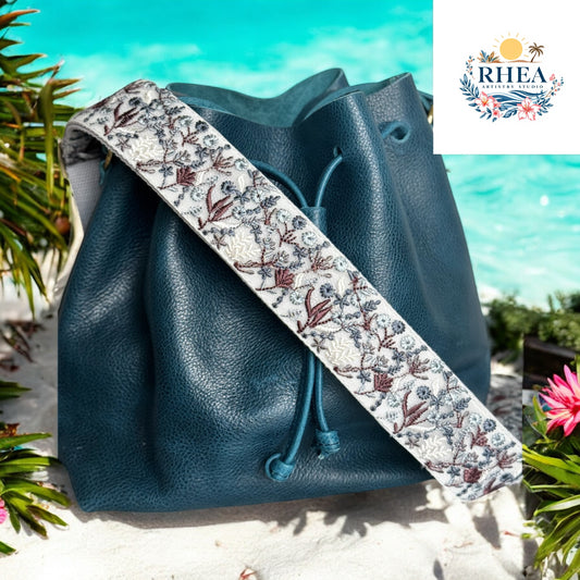 A 2-inch cream strap with blue and brown embroidered foliage, styled on a navy leather bucket bag.