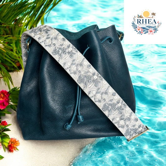 A neutral floral embroidered bag strap in soft silver and gray tones, displayed on a dark teal leather bucket bag.