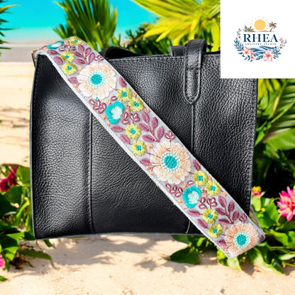 A black handbag with a floral strap featuring aqua, coral, and lavender embroidery. Set in a beach