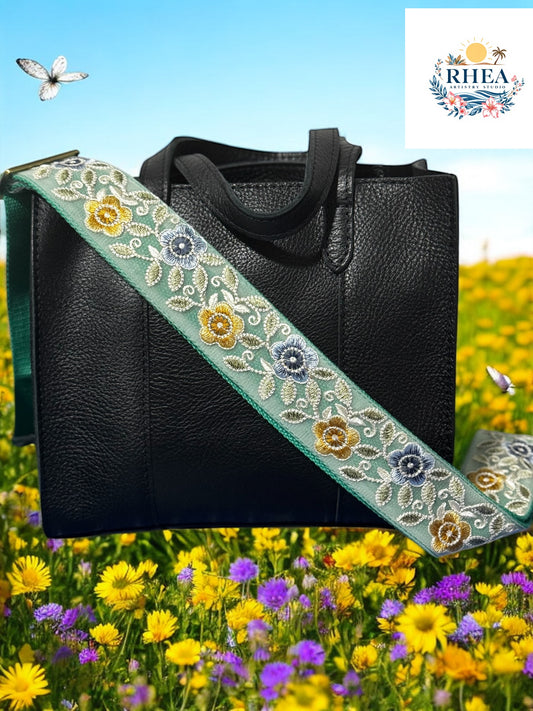 Black leather handbag with a handmade embroidered floral crossbody strap featuring yellow, blue flowers with green and yellow vines, displayed on a garden.