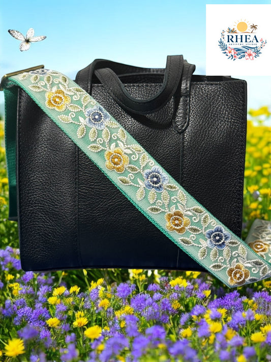 Black leather handbag with a handmade embroidered floral crossbody strap on a sea foam green cotton webbing featuring yellow, blue flowers with green and yellow vines, displayed on a garden.