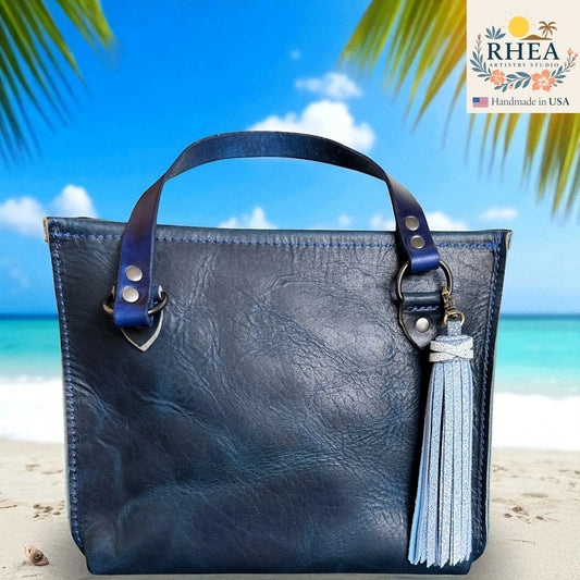 Blue leather handbag with a tassel on a beach background, featuring the RHEA brand.