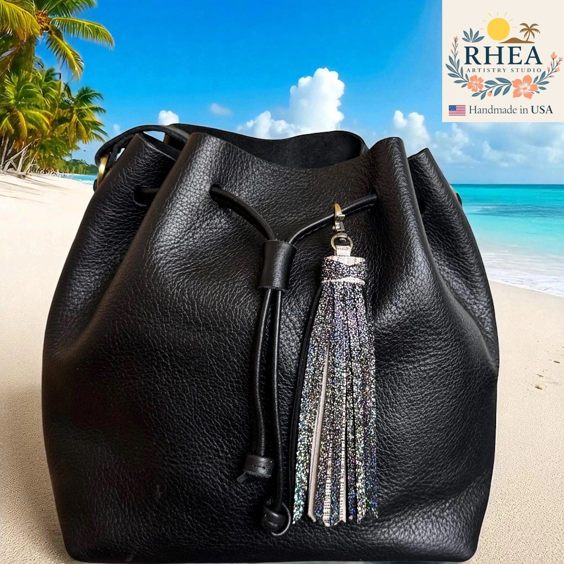 Black leather bucket bag with a tassel on a beach background, featuring the RHEEA brand.