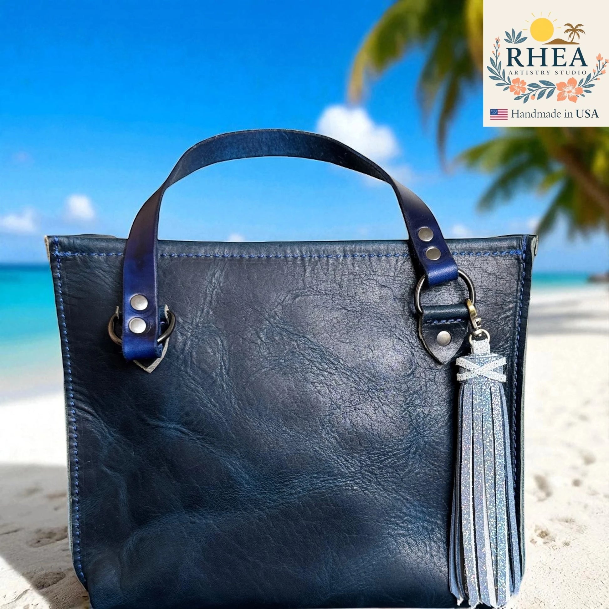Blue leather handbag with tassel on a beach, featuring the RHEA Artistry Studio logo.