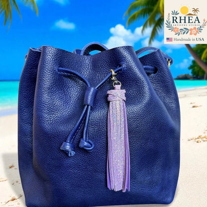 Blue leather bag with a purple tassel on a beach background, featuring the RHEA Artistry Studio logo.