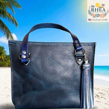 Blue leather handbag with tassel on a beach background, featuring the RHEA brand.