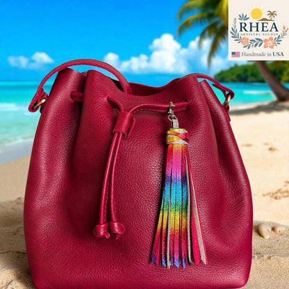 Red leather bucket bag with a colorful tassel on a beach background, featuring the RHEA brand.
