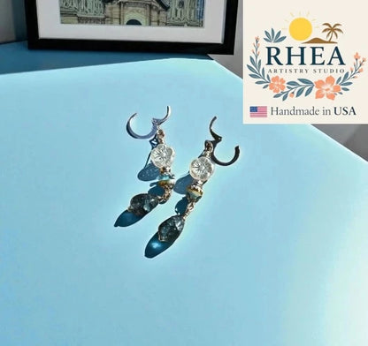 Decorative earrings on a light blue surface with RHEA Artistry Studio logo.