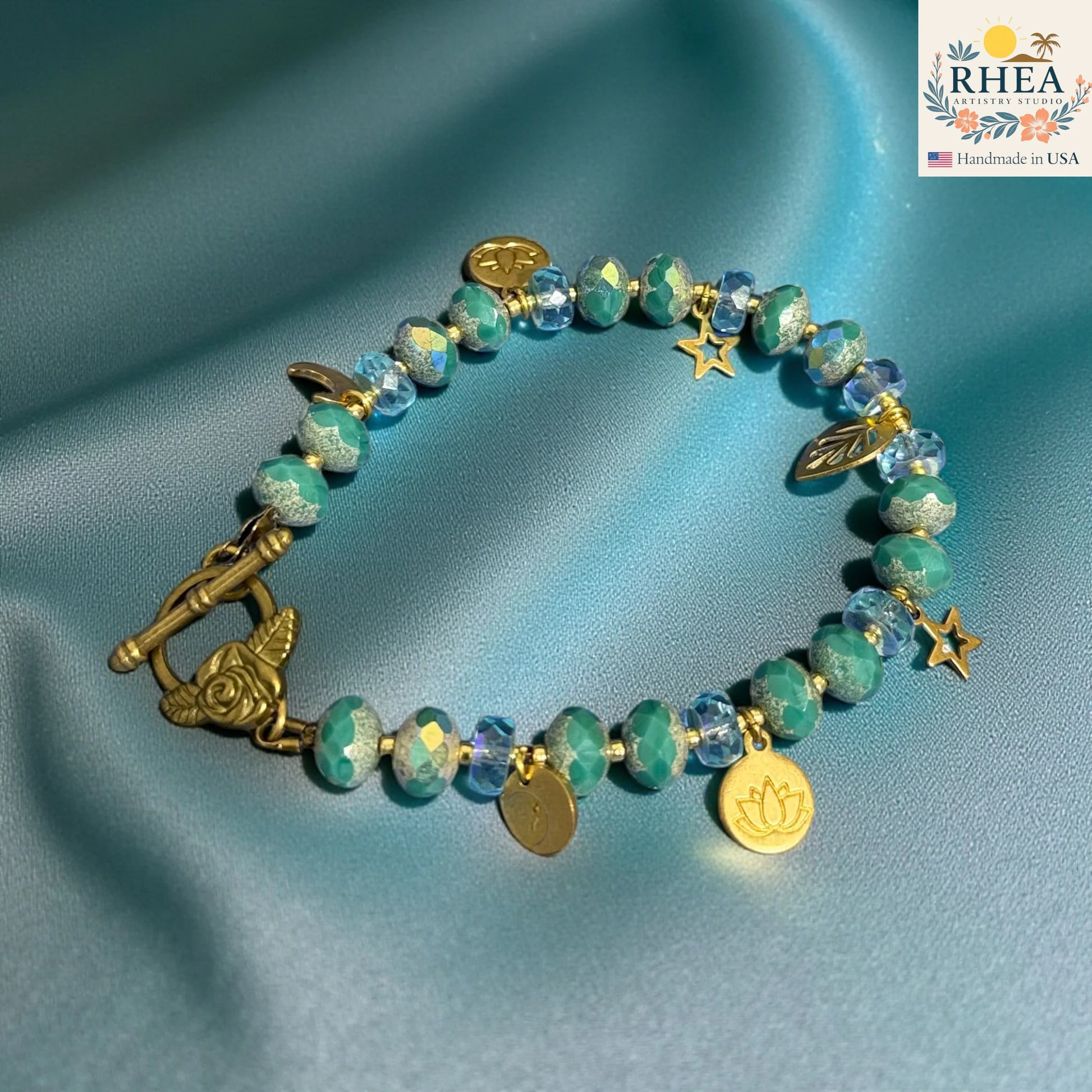 Turquoise beaded bracelet with gold charms on a blue fabric background, featuring the RHEA brand.