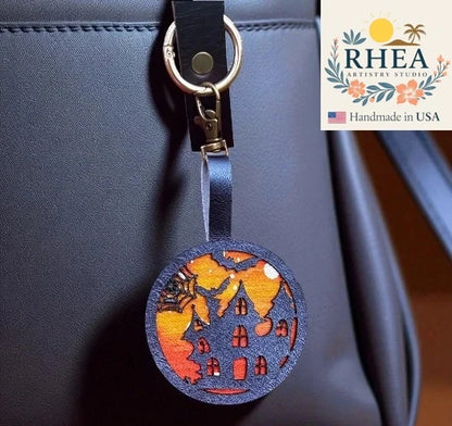 Decorative leather purse charm with house design on a blue bag, featuring RHEA Artistry Studio logo.