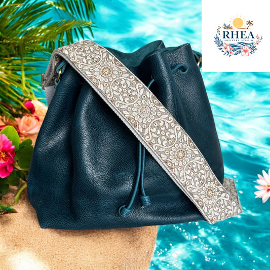 2-inch wide artisan strap with champagne gold and ivory mandala floral embroidery, displayed on a navy leather bag near turquoise water.