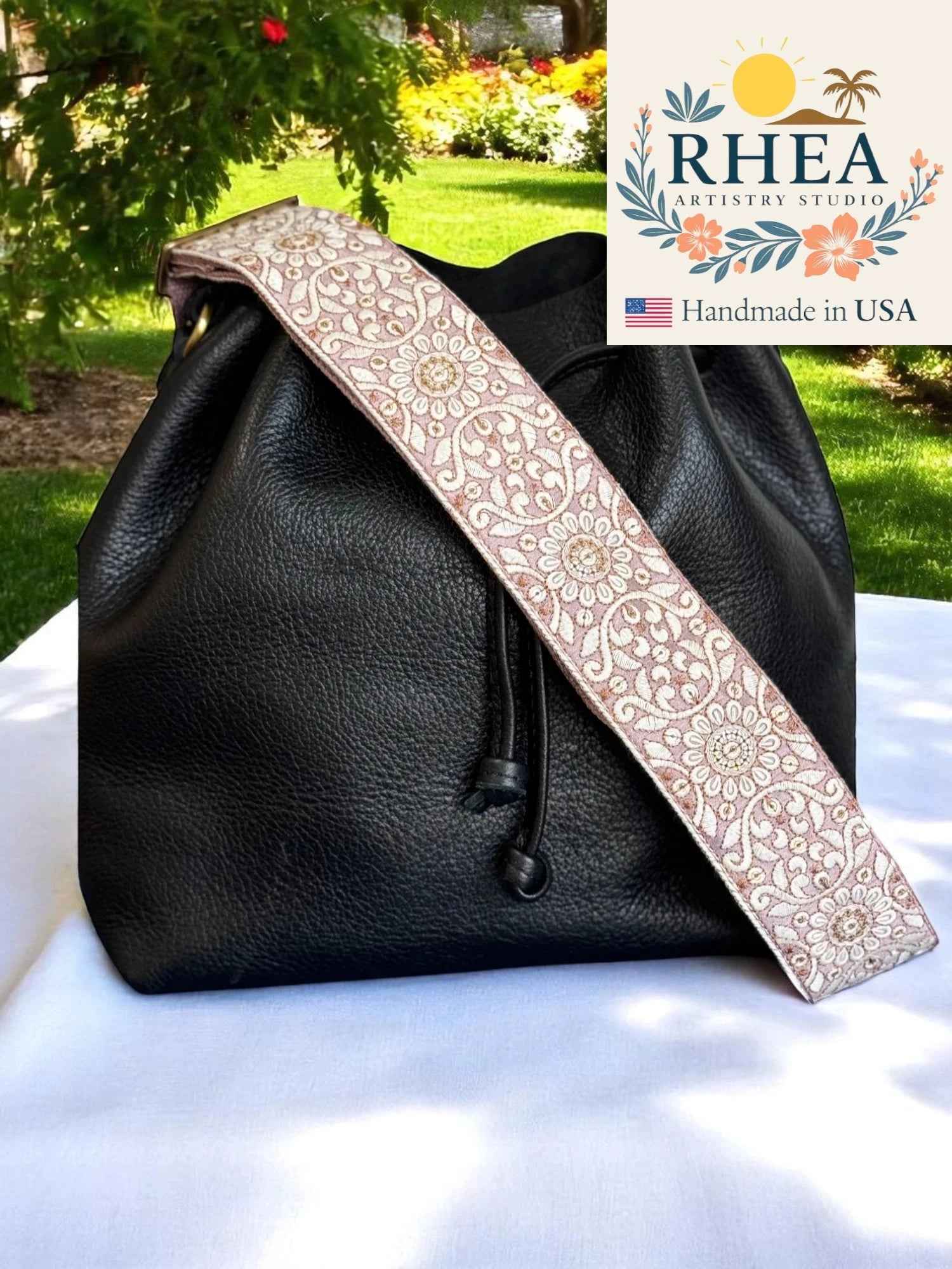 Black leather bag with a decorative purse strap on a white surface outdoors, featuring the RHEA Artisan Studio logo.