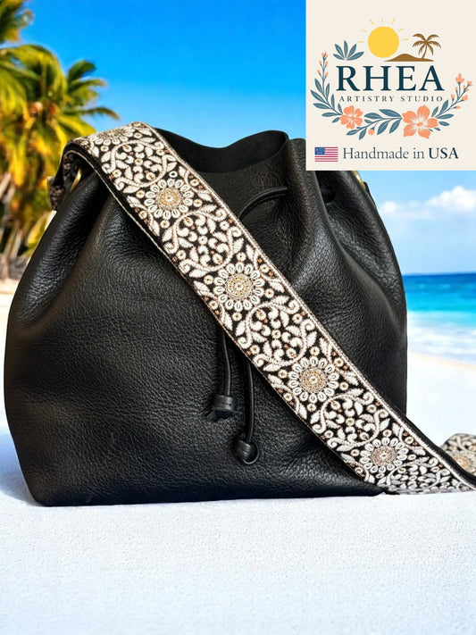 Black leather bag with decorative purse strap on a beach, featuring RHEA Artists Studio logo.