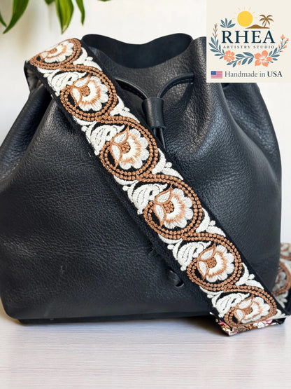 Black leather bag with decorative purse strap featuring RHEA Artisans Studio branding.
