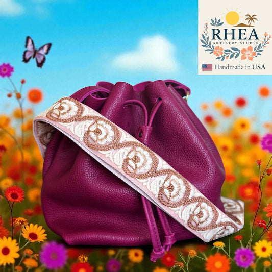 Purple drawstring bag with decorative purse strap in a field of flowers and butterflies, featuring the RHEA Artistry Studio logo.