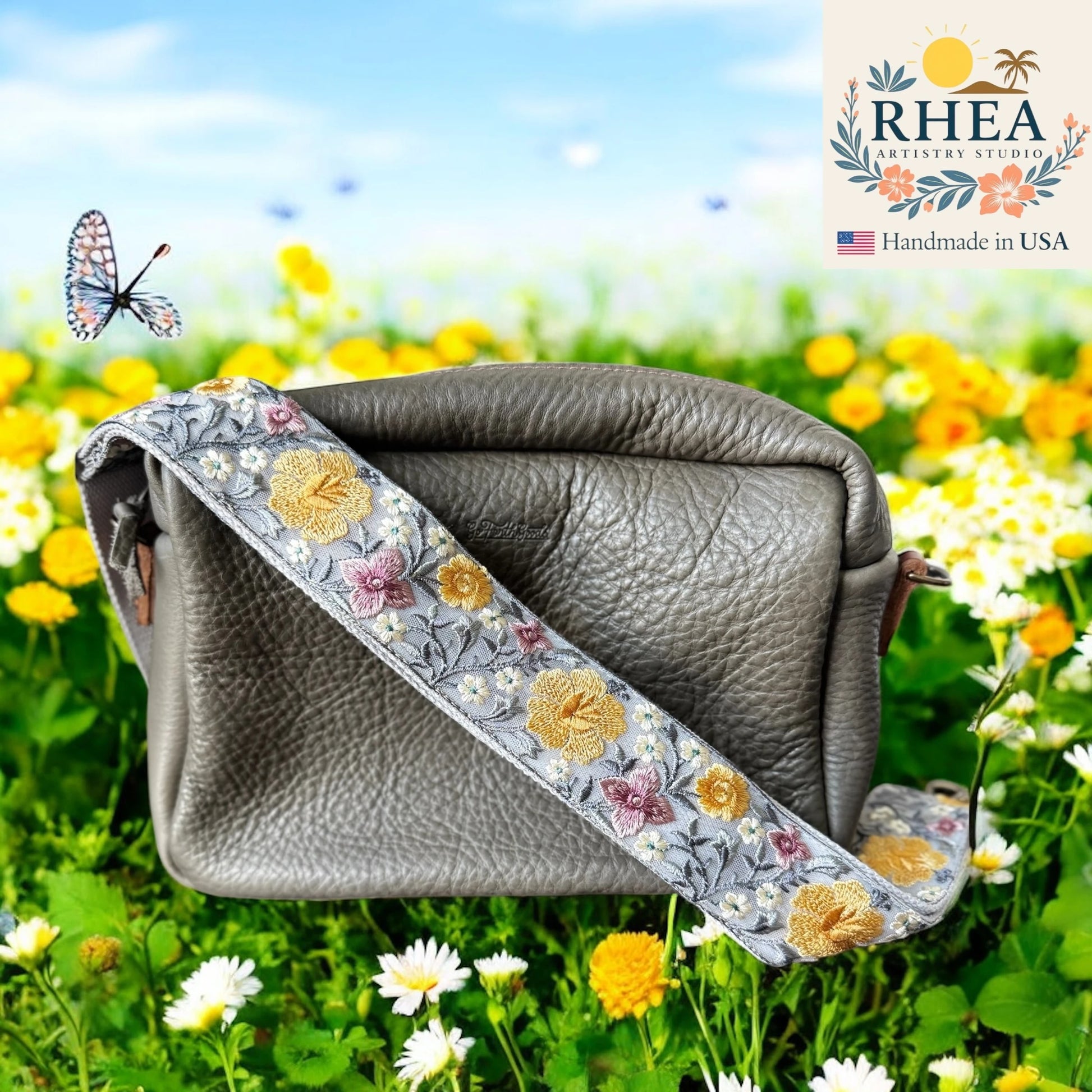 Gray leather bag with floral purse strap on a grassy field with flowers, RHEA brand visible