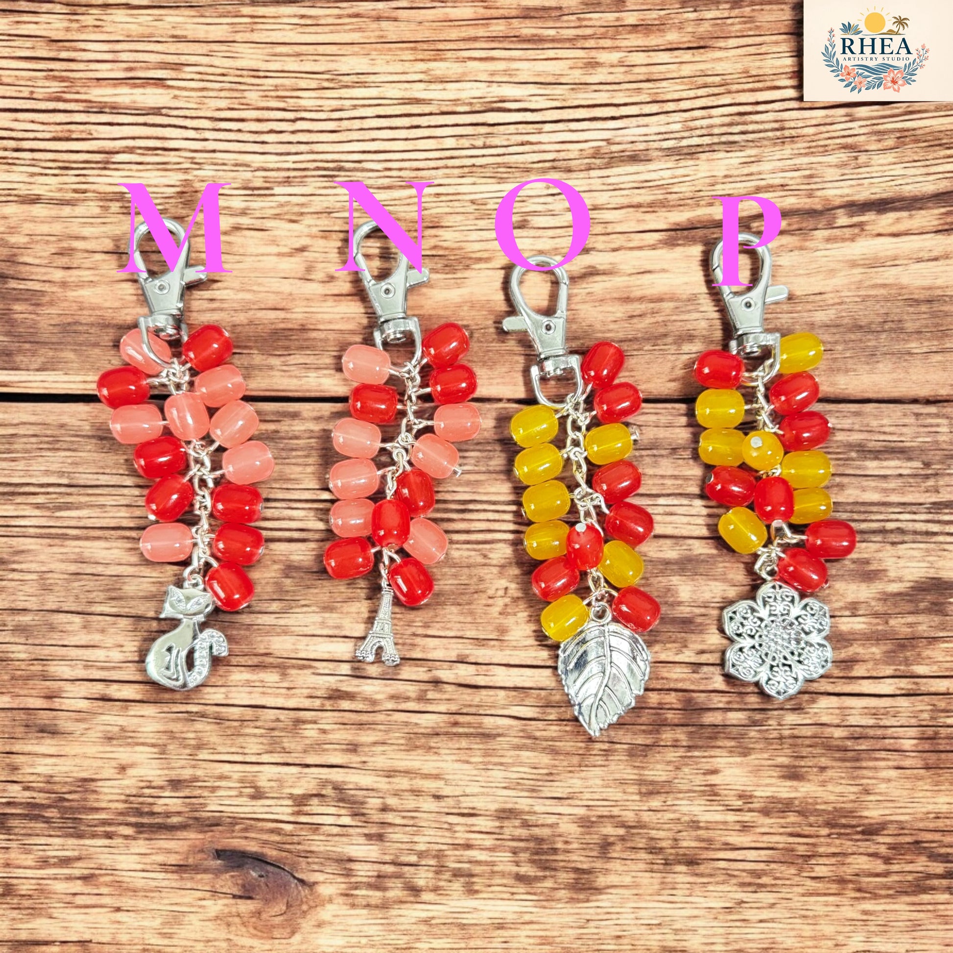 Four colorful keychains with letter charms on a wooden surface, branded 'RHEA'.