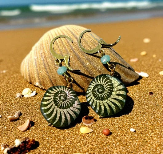 Pair of earrings with green spiral designs on a beach with shells and sand.