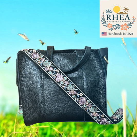 Black leather handbag with floral strap against a natural background, featuring the RHEA Artistry Studio logo.