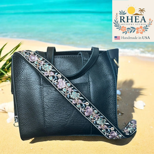 Black handbag with floral strap on a beach, featuring RHEA Artistry Studio logo.