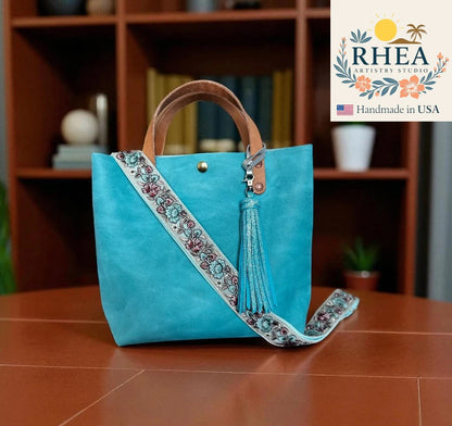 Blue handbag with floral embroidery and tassel on a wooden surface, with RHEA Artistry Studio logo in the corner.