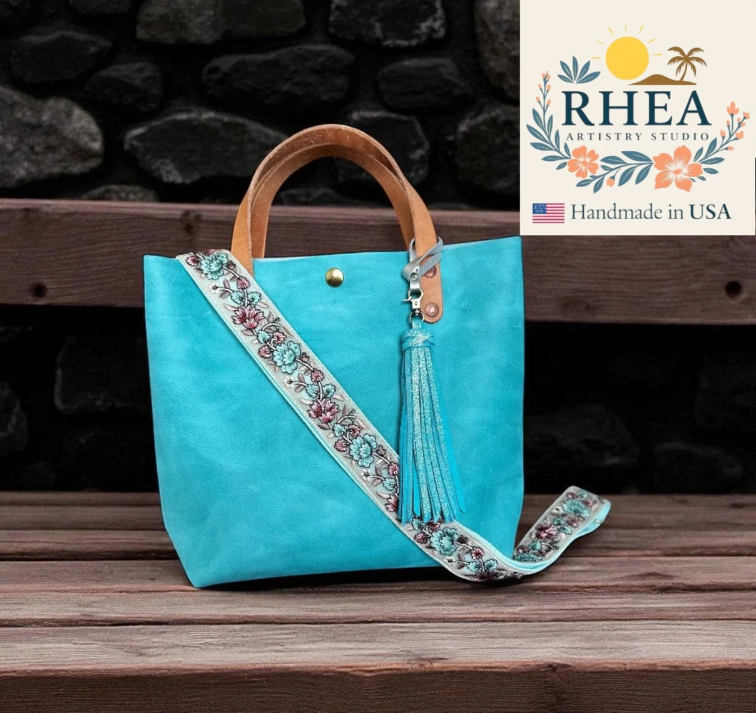 Turquoise handbag with decorative strap and tassel on a wooden surface, branded 'RHEA Artistry Studio'.