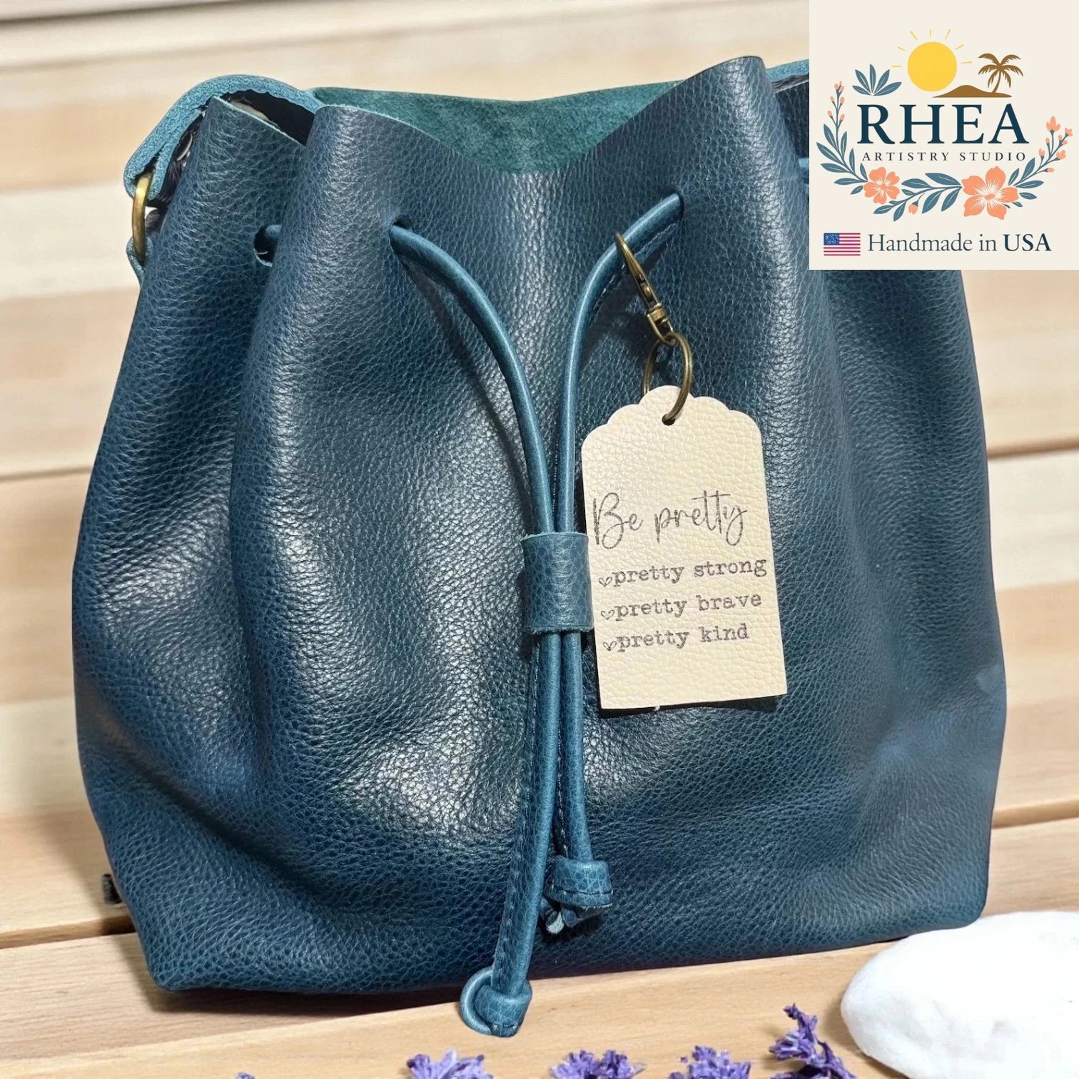 Blue leather bag with a leather tag on a wooden surface, featuring the RHEA brand.