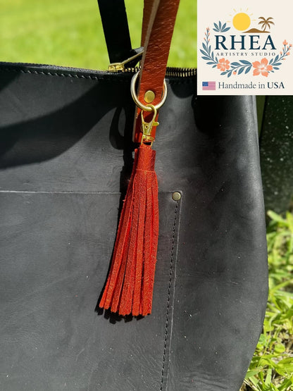 Black leather bag with a orange tassel and 'RHEA' branding in the corner.