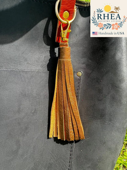bronze leather tassel keychain attached to a black leather bag with RHEEA Artisan Studio branding.