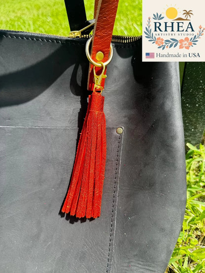 Black leather bag with a orange tassel on a grassy background, featuring the RHEA Artistry Studio logo.