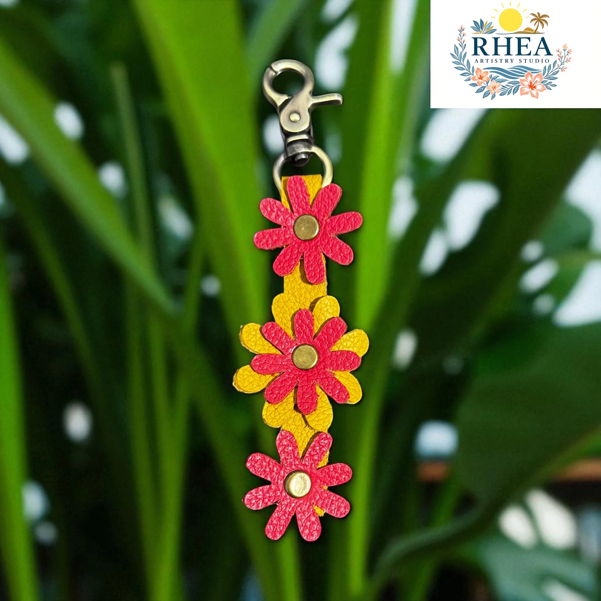 Floral keychain with pink and yellow flowers on a green leafy background, featuring the RHEA brand logo.