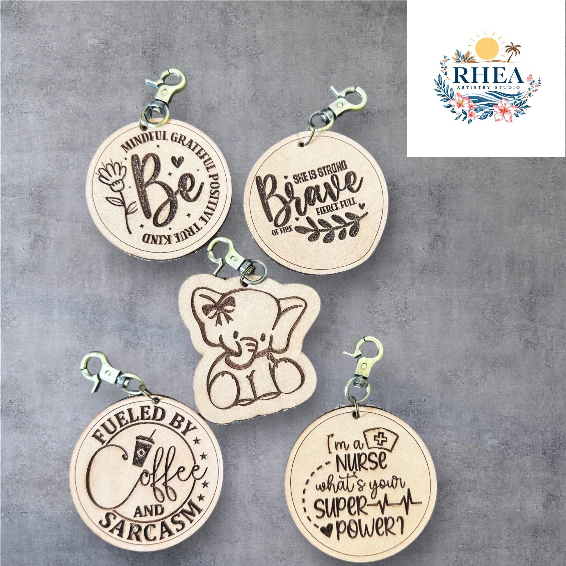 Five wooden keychains with engraved text on a gray background, featuring the brand 'RHEA'.