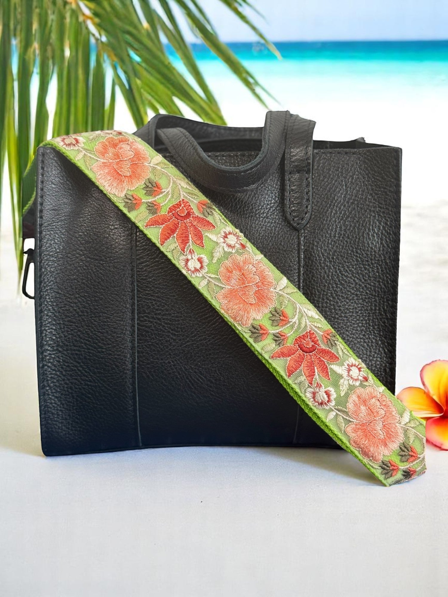 A black purse with a pink and green floral embroidered strap placed on a surface with a field of flowers in the background.