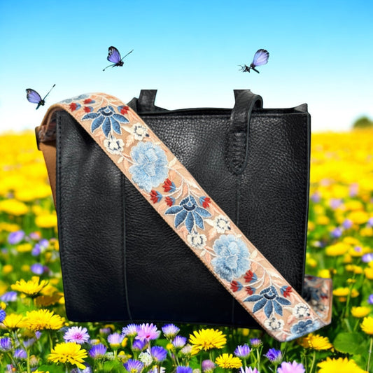 Black leather handbag with a wide light orange embroidered strap featuring blue flowers, navy leaves, and red accents, set against a vibrant field of yellow and purple wildflowers — Rose Bloom Collection by Rhea Artistry Studio.