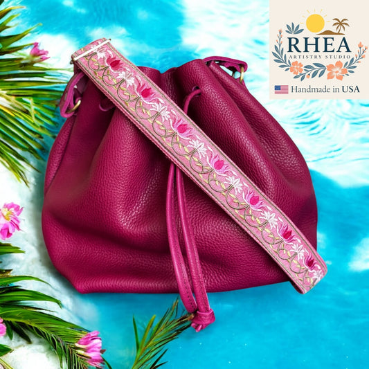 Pink leather drawstring bag with floral purse strap on a blue background, featuring RHEA Artistry Studio logo.