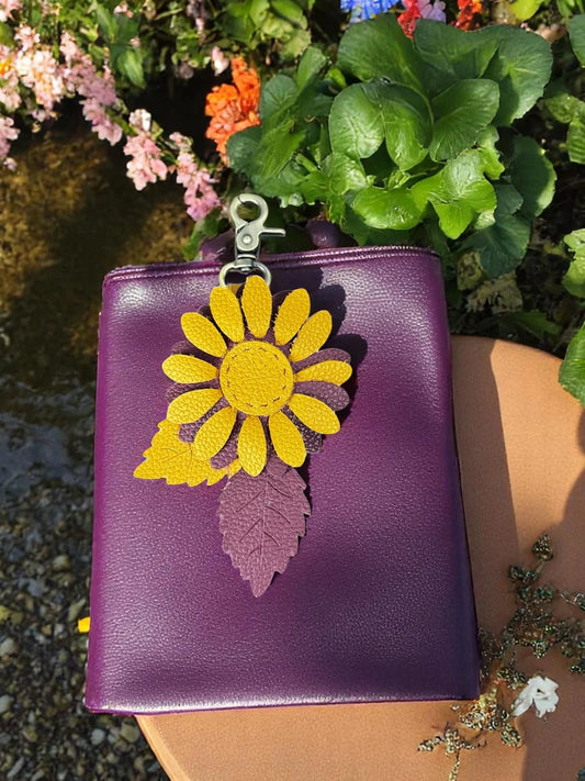 Hand-sewn leather daisy charm in marigold and plum with brass clasp and textured leaves in the garden